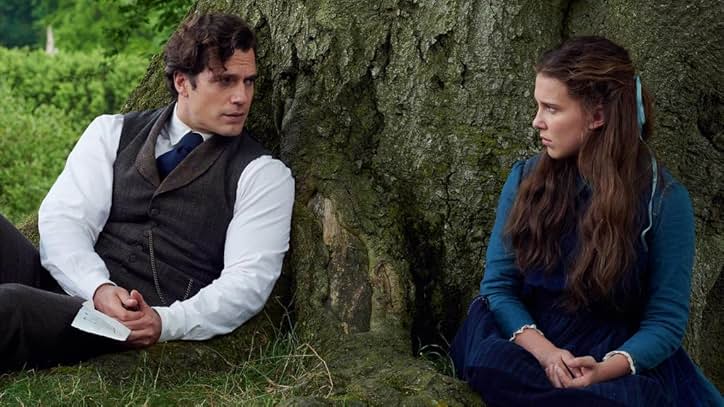 A still from Enola Holmes. A man in a gray vest, white shirt, and blue tie leans against a tree and looks to his right. Looking back at him is a girl wearing a blue dress and a blue hair ribbon.