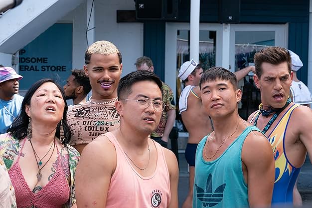 A still from Fire Island. A woman in a pink crochet top, and three men wearing tan, pin, blue, and yellow and blue tops look off in the distance with a mix of amusement, confusion, and shock.
