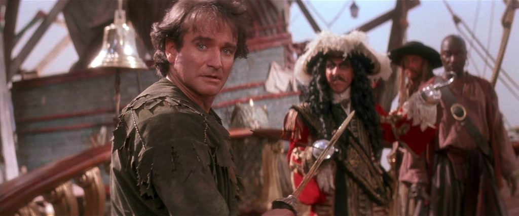 A still from Hook. A man wearing a green leafy costume stands on a pirate ship holding a short sword and looking over his shoulder. Behind him is a pirate wearing a feathery hat, curly black wig, and red jacket. He holds a sword and has a hook for a hand. Behind him are two other pirates.