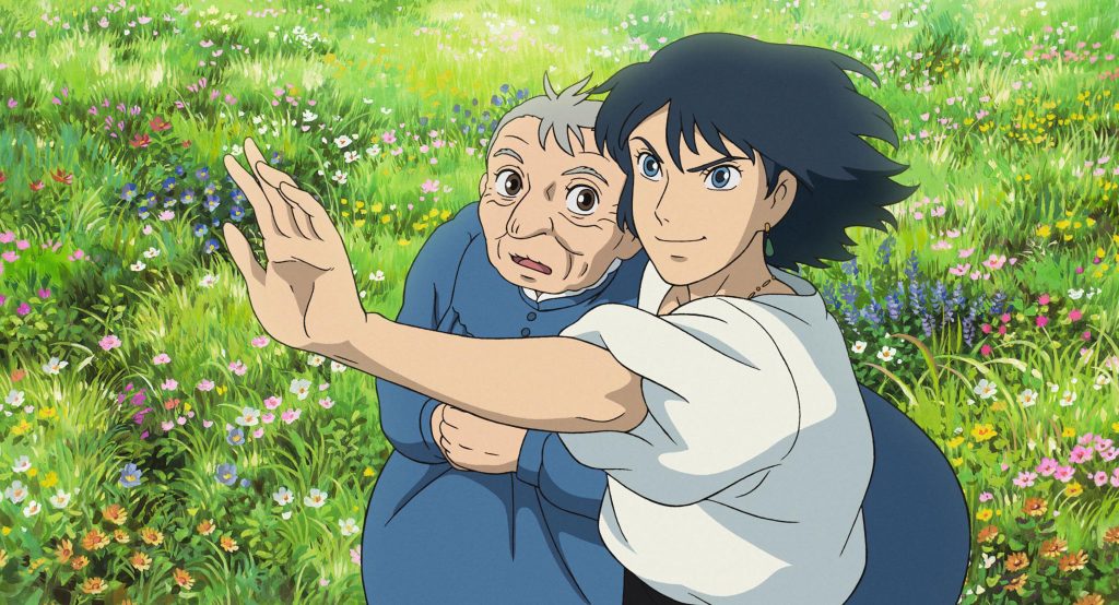 A still from Howl's Moving Castle. A field full of flowers. A man with black hair, a white blouse, and green earrings holds his hand out in front of him and tightens his face in concentration. Beside him, an old woman in a blue dress looks on in wonder.