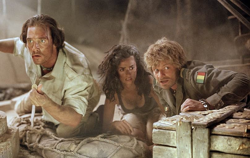 A still from Sahara. A man wearing white shirt and khaki pants, a woman wearing a black tank top and khaki pants, and a man wearing a brown military uniform crouch inside a ship's hull. They are covered in dust and surrounded by boxes, ropes, and gold coins.