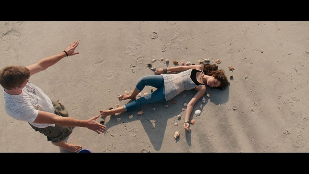 A still from The Last Song. A young woman lies on the beach surrounded by seashells. She wears a white lace top and jeans. Above her is a young man in a white t-shirt and green shorts holding his arms out in admiration.