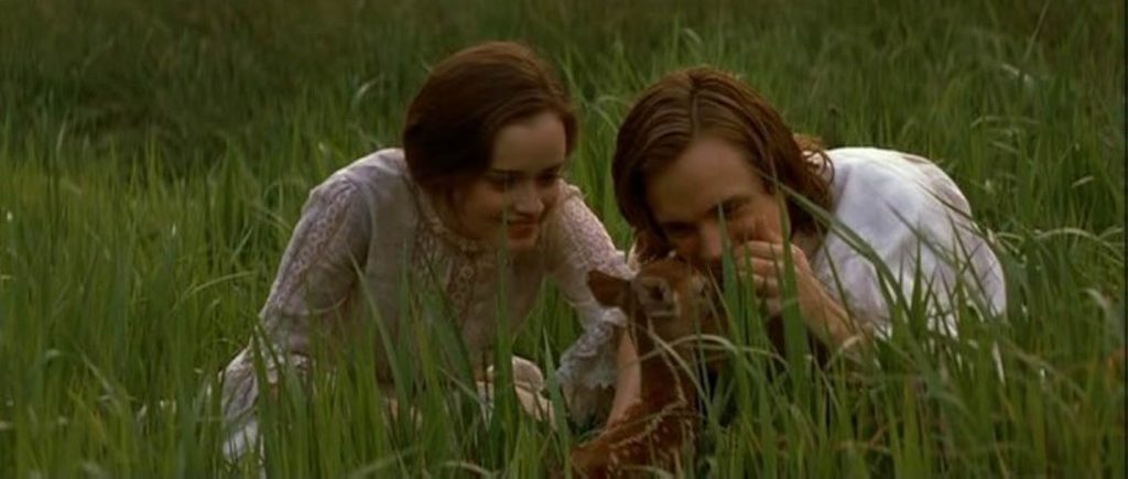 A still from Tuck Everlasting. A young woman in a white lacy dress crouches in a grassy field beside a young man in a white shirt. The man reaches forward to pet a baby deer.