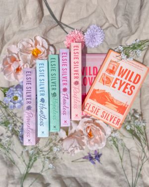 A flatlay featuring seven of Elsie Silver's books laid on a rumpled beige sheet. Five of them are arranged with their spines facing out, from left to right: Powerless, Heartless, Hopeless, Flawless, and Reckless. Two of them are laid flat, from top to bottom: Wild Eyes and Wild Love. The books are surrounded by pink and light purple flowers.