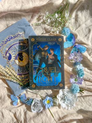 A flatlay featuring the novel Cruel Is the Light by Sophie Clark atop a rumpled white sheet. To match the cover's blue background, various blue flower and butterfly decorations have been arranged to frame the book, along with a blue pouch featuring an embroidered celestial design.