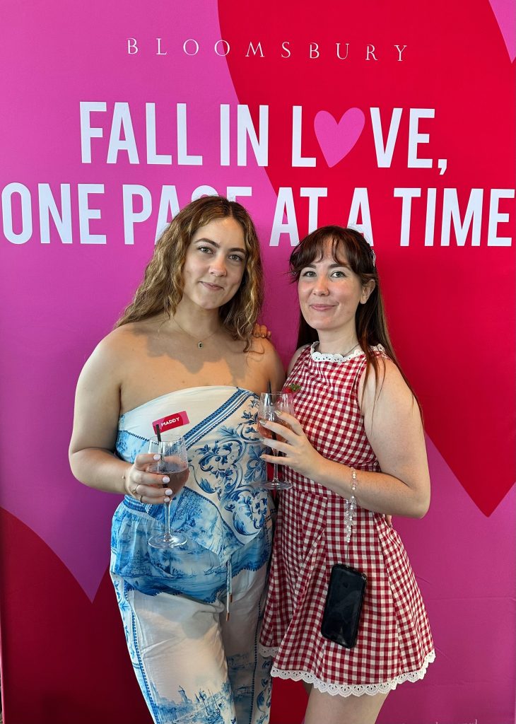 Bookstagrammers Maddy and Allie standing in front of a pink backdrop with large red hearts reading "Bloomsbury: Fall in Love, One Page at a Time." Maddy has long, wavy dark brown/blonde hair wears a strapless white top with a blue pattern and matching white pants with a blue pattern. Allie has brown hair tied half up, and wears a red checkered dress. She has a phone hanging off a strap around her wrist. Each sister is holding a glass of red wine.