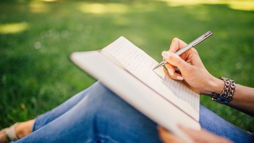 A manicured hand holds a silver pen and writes in a journal outside.