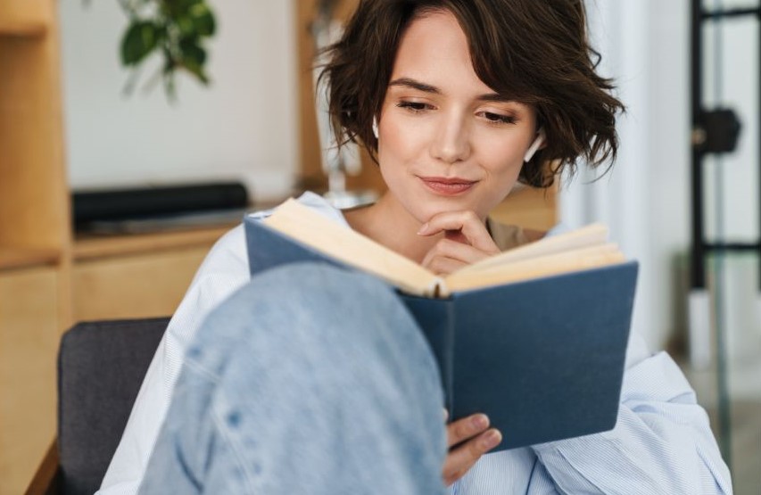 A woman reading a dark blue book with a title on it. She's sitting with her knees propped up, and she's faintly smiling while reading.