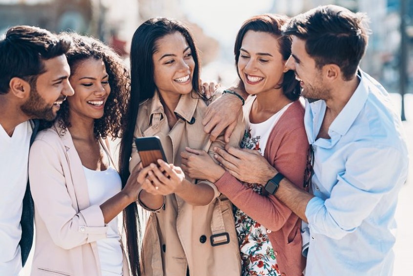 Five people of different skin tones, genders, and hair textures looking at a phone held by the person in the middle. They're all smiling and/or laughing.