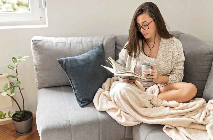 A woman sitting on the couch reading a book. A blanket is thrown over one leg, and she has a drink held in one hand.
