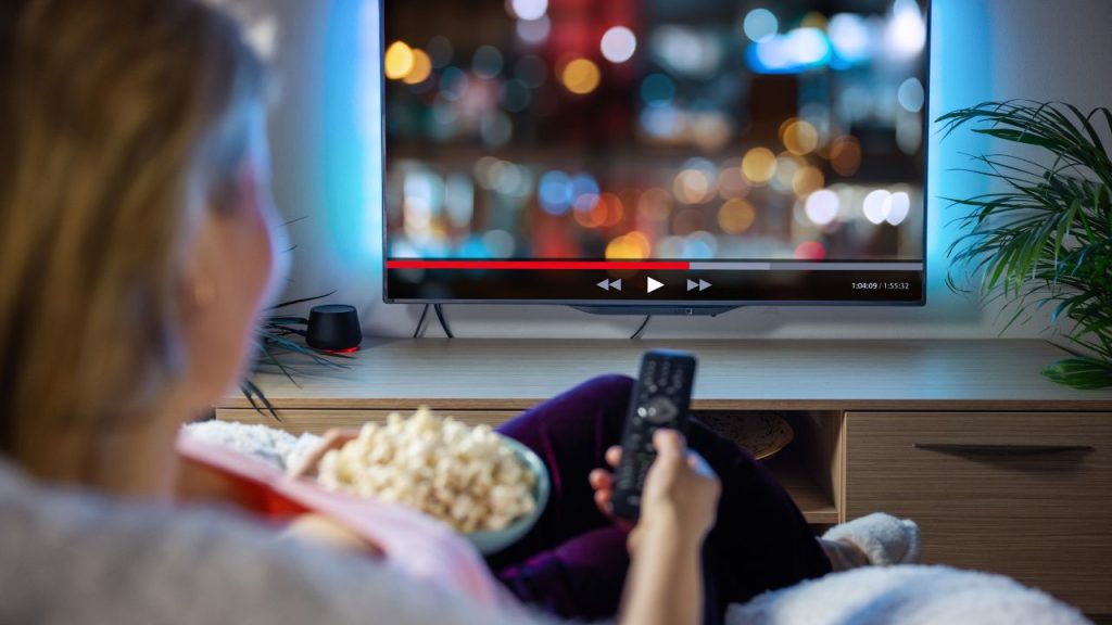 A woman holds a bowl of popcorn and a remote pointing at a paused TV screen.