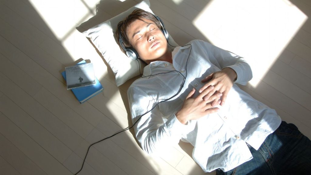 A man laying on the floor with a journal next to him, his eyes closed, and his head on a pillow, listening to music on headphones.