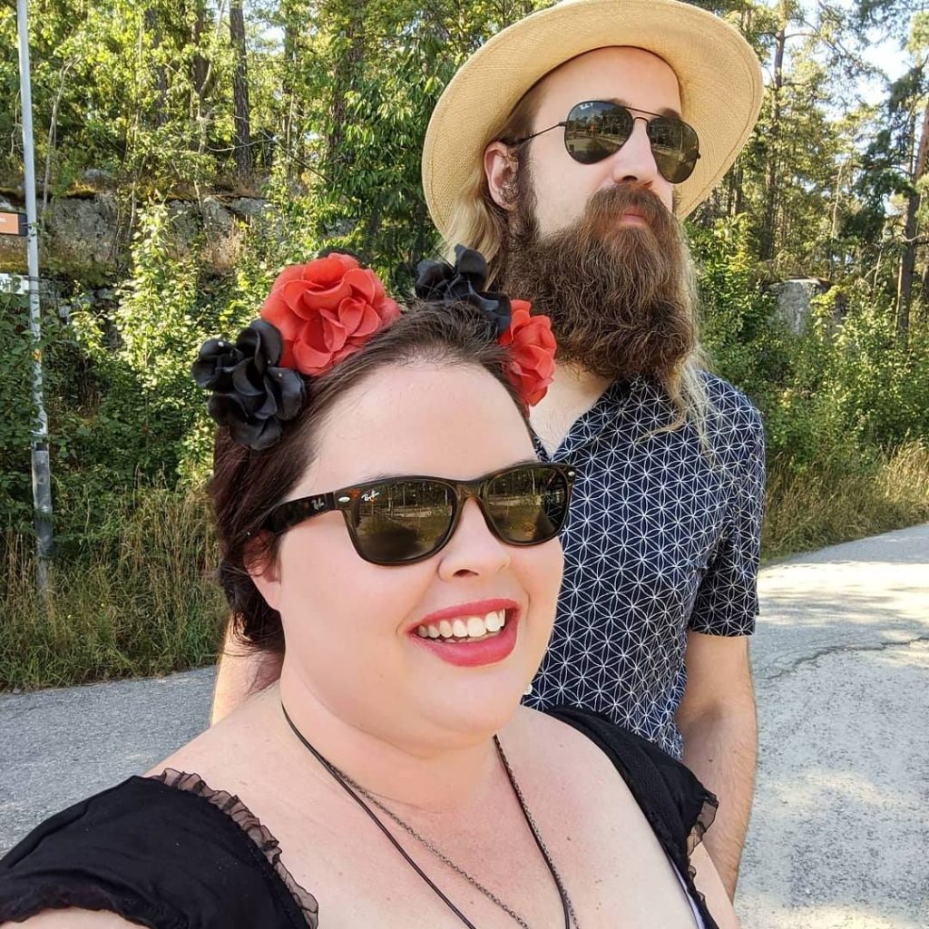 A man and a woman stand outside against a green woodsy background. The man is wearing a beige straw hat and sunglasses. The woman is wearing a red and black flower crown and sunglasses.