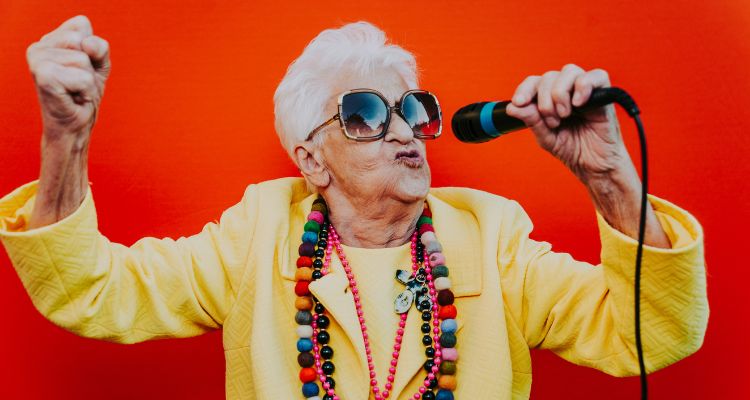 Older woman with short white hair is wearing large sunglasses and a yellow blazer. She is holding a mic in one hand and raising her other hand in a fist.