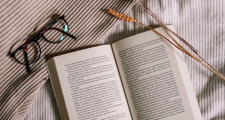 Open books laying on a tan blanket with glasses above it and a piece of hay on one of the corners