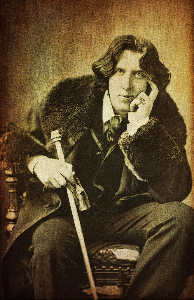 Oscar Wilde poses with one hand holding his face and another around his can while staring into the camera.