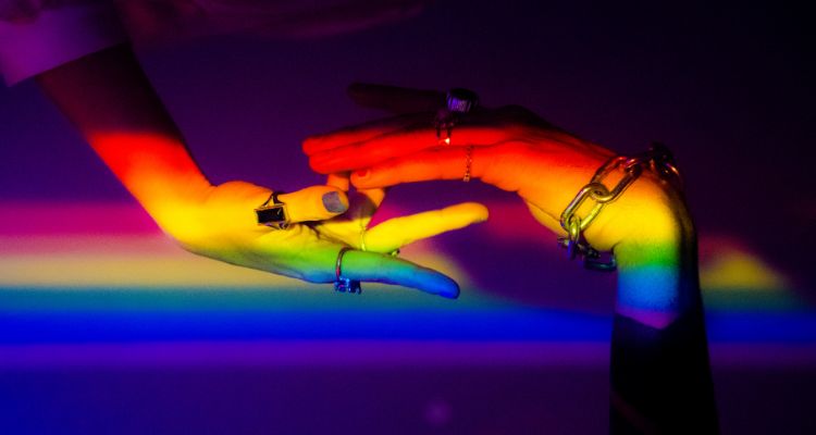 Two feminine presenting hands, hovering over eachother. Both wear rings and bracelets and have painted nails. There is a light rainbow covering the hands and the background is pitch black.