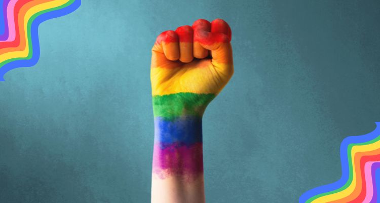 Image of a dark blue background. A hand is in the center, fisted and painted rainbow to represent Pride. There are swirly rainbows in two of the corners.