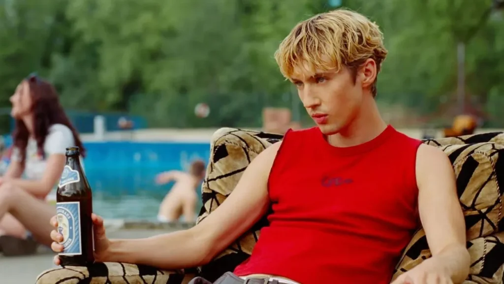 Still from Rush by Troye Sivan Music Video, depicting Troye Sivan in a red tank top sitting on a loveseat holding a beer. The background behind him is blurred but there seems to be people in and around a pool.