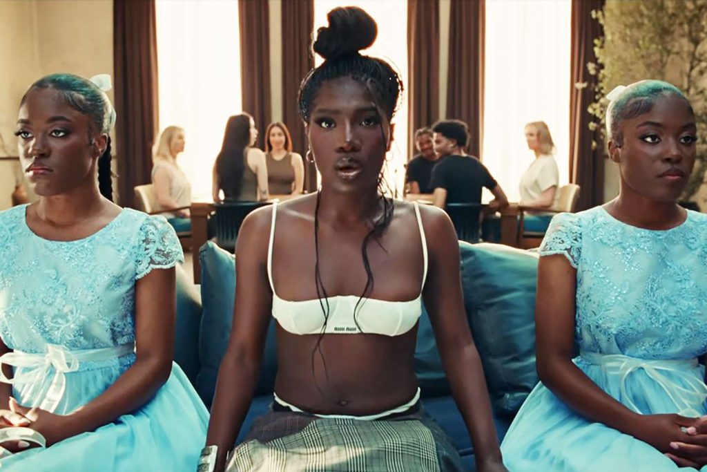 Still of Anxiety by Doechii Music video, depicting Doechii looking at the camera while sitting on a couch next to two women looking ahead. Behind them, six people sit around a table.