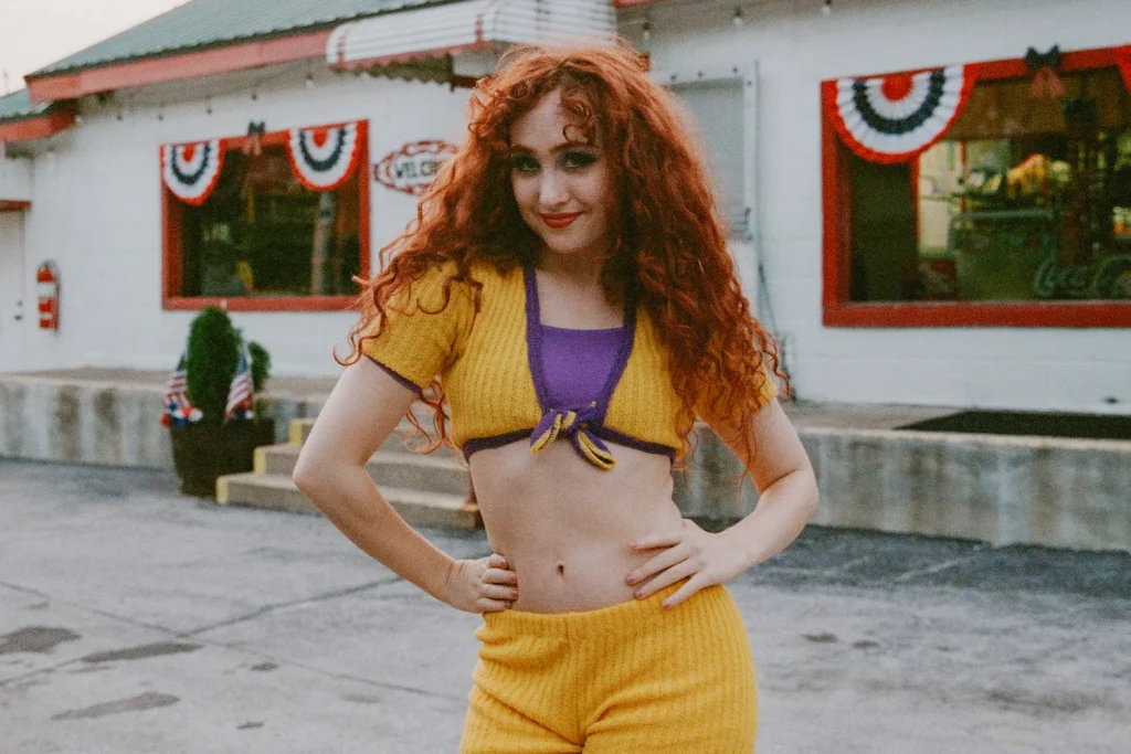Still from HOT TO GO by Chappell Roan music video, depicting Chappell Roan in a knit yellow and purple top and yellow bottoms. She is standing in the entrance of a restaurant.