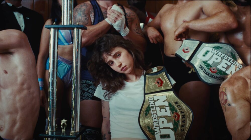 Still from Juna by Clairo music video, depicting Clairo with a wrestling belt over her shoulder, surrounded by male wrestlers.