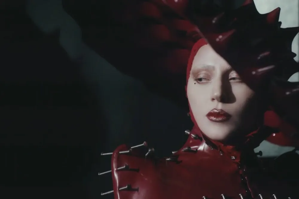 Still from Abracadabra by Lady Gaga Music Video, depicting Lady Gaga in a red latex suit and hat with metal nails. She is looking to the side and the background is dark.