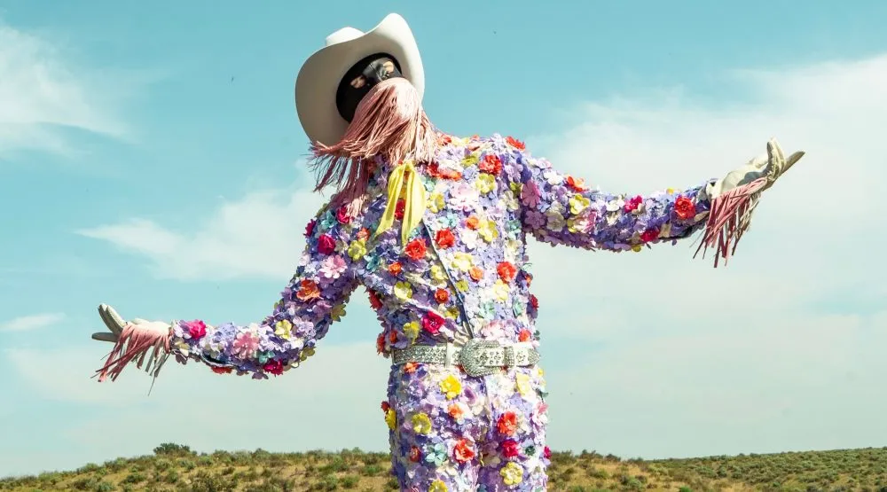 Still Image from Summertime by Orville Peck music video, depicting Orville Peck in a black mask with pink fringe, white cowboy hat, and floral suit. The background features a grassy hill and blue sky.