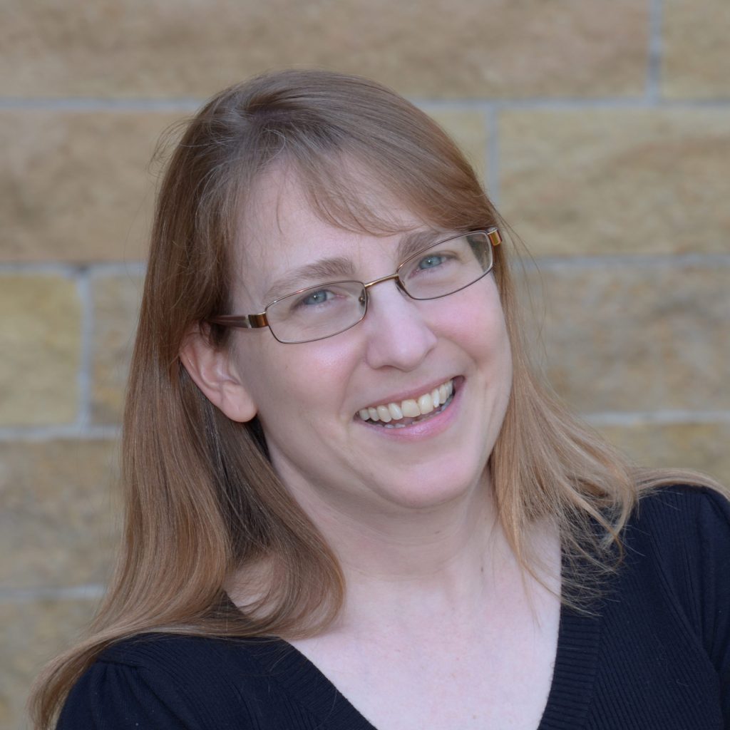 H.M. Bouwman, a smiling light-skinned woman with long, straight light brown hair and blue eyes, wearing glasses and a black v-neck sweater, standing outdoors near a tan brick wall.