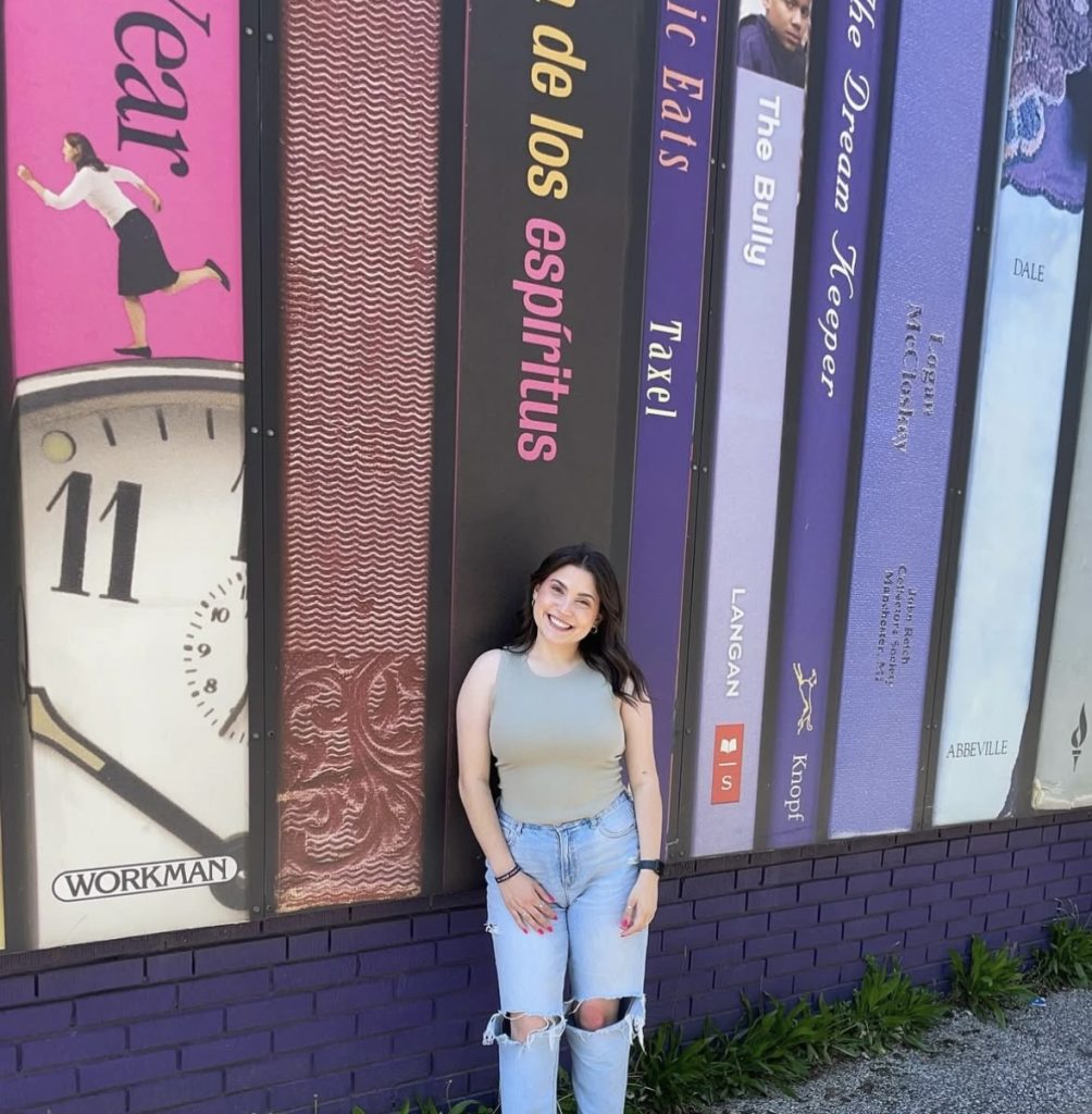 Bookstagrammer Lindsey outside smiling in ripped jeans and a tan tank top in front of a wall decorated with book bindings.