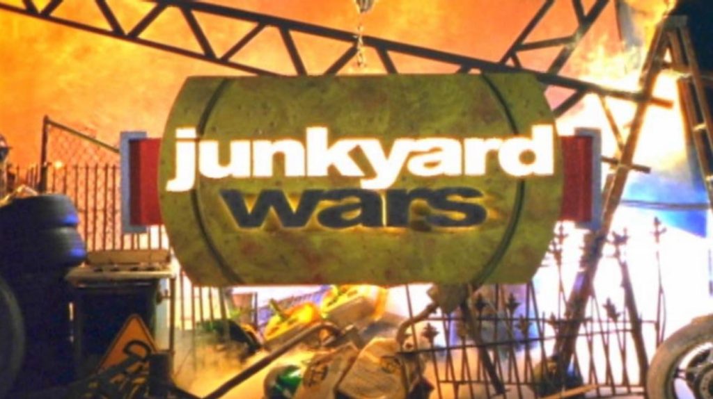 Screenshot of title card from television show Junkyard Wars, featuring the title on a metal plate against a background of various scrap metal and discarded items.