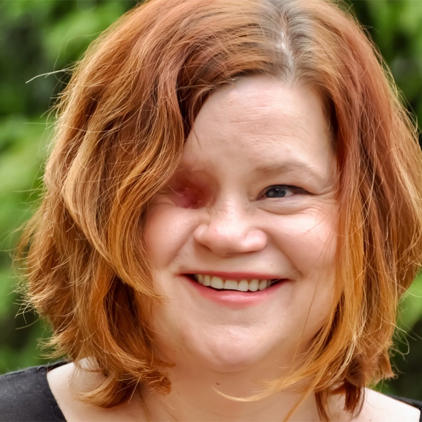 Author Katie Mazeika, a light-skinned woman with chin-length, straight red hair and one blue eye, wearing a black blouse and standing outside in a green, natural area. She is missing one eye.