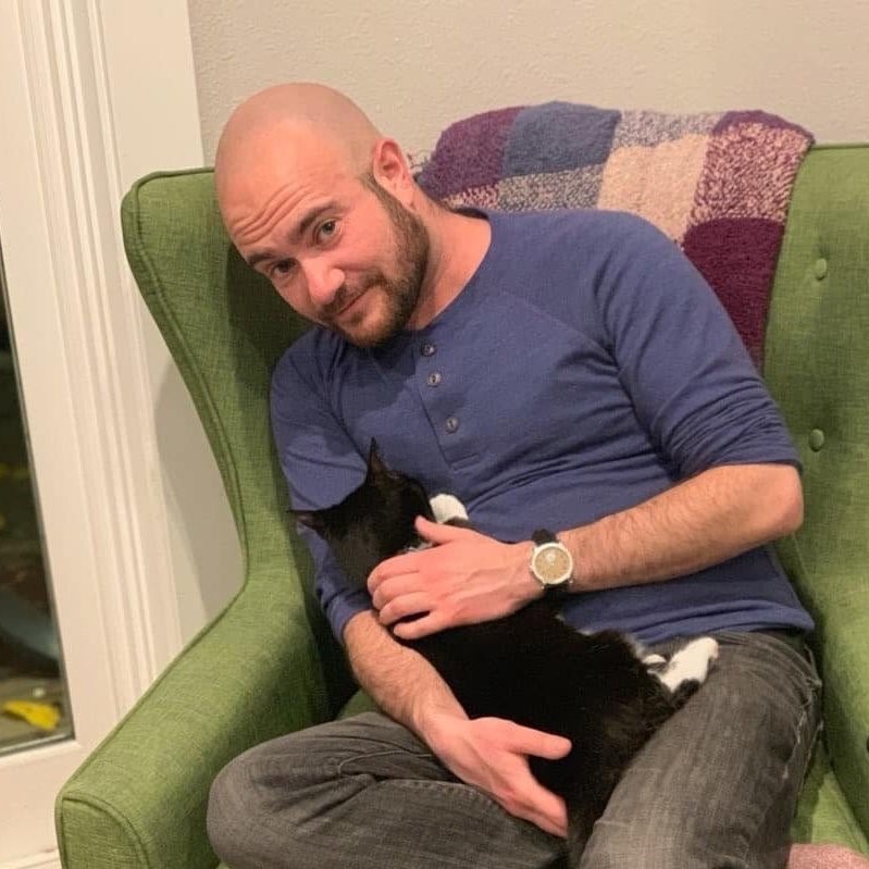 Kyle Lukoff, a light-skinned man with a brunette beard and shaved head, wearing a blue long sleeved henley shirt and sitting in a green armchair with a black and white cat in his lap.