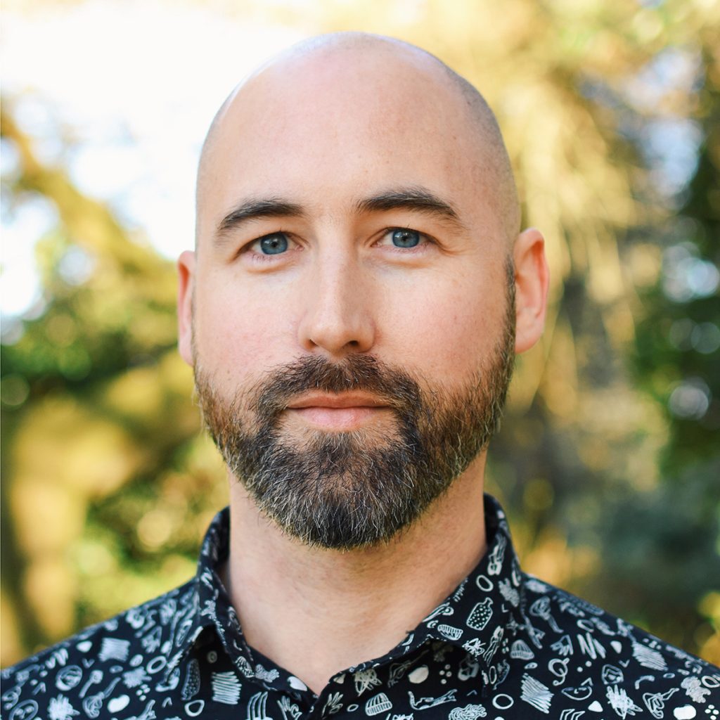 Levi Hastings, a light-skinned man with a brunette beard, a shaved head, and blue eyes, wearing a black and white collared shirt and standing in an outdoor wooded area.