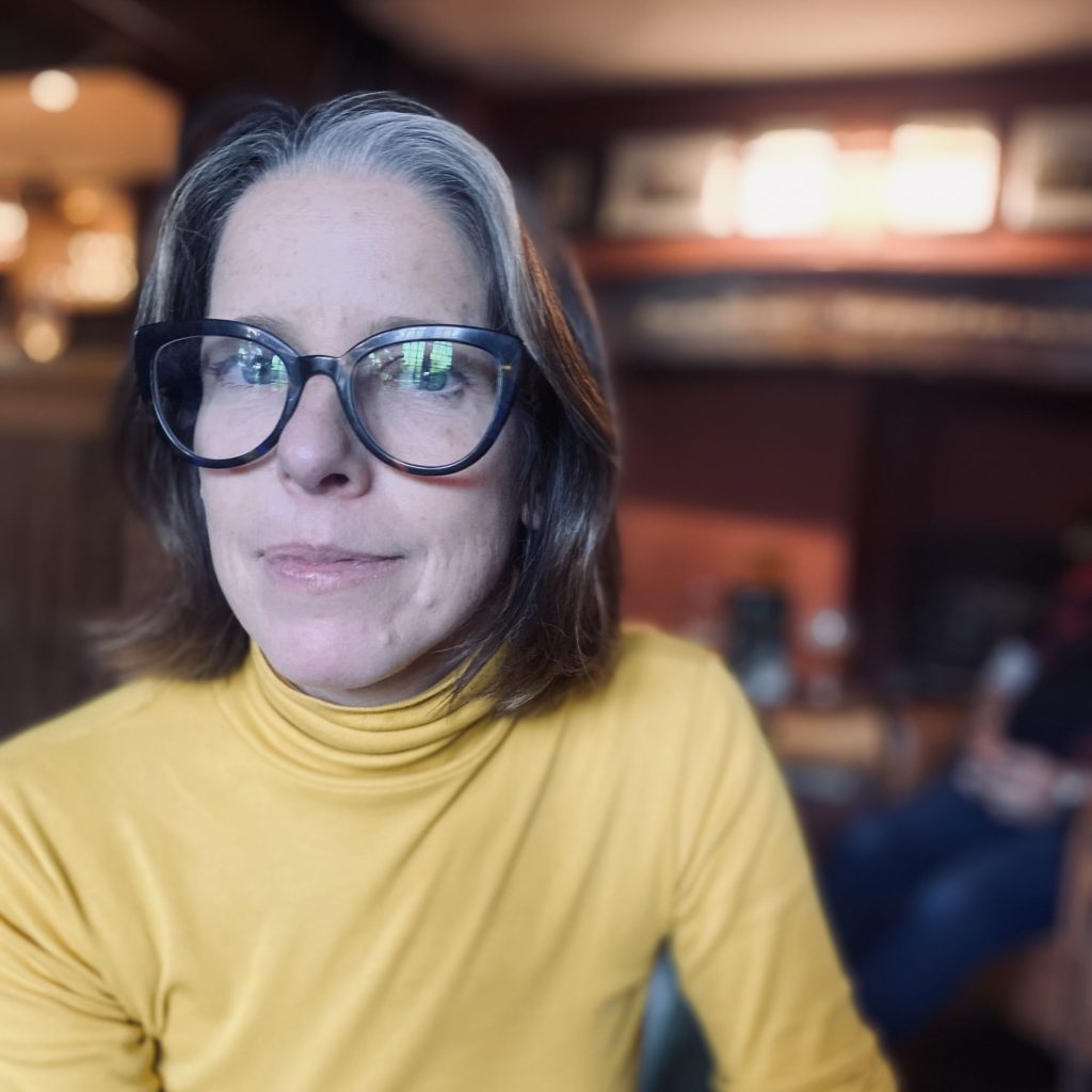Madeleine Rogers, a smiling light-skinned woman with shoulder-length, straight gray and brown hair and blue eyes, wearing glasses and a yellow turtleneck, sitting in an interior space with dark red walls.