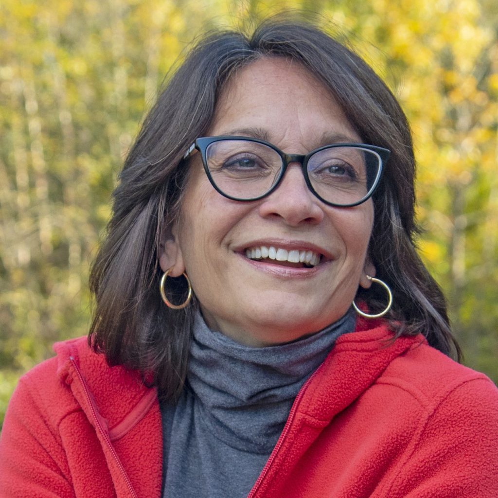 Meg Medina, a smiling tan-skinned woman with straight, brown, shoulder-length hair and brown eyes, wearing glasses, gold hoop earrings, a gray turtleneck and a red fleece, in an outdoor area with trees visible in the background.