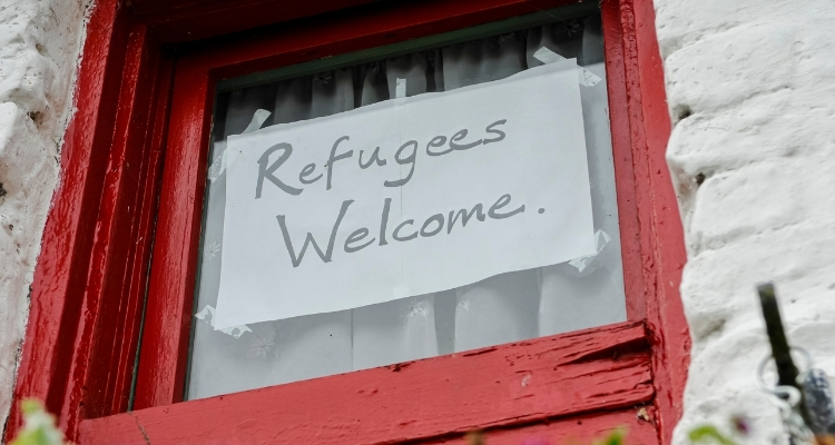 A paper sign taped to the inside of a red-framed window reads "Refugees Welcome."