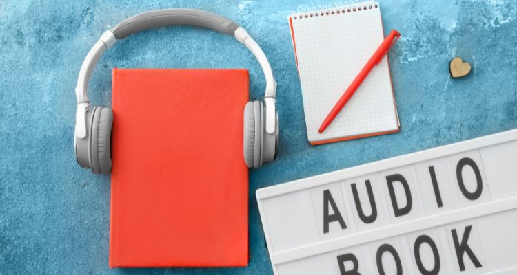 The image depicts a red book with headphones around it with display text, "Audiobook" and a notepad on a light blue surface.