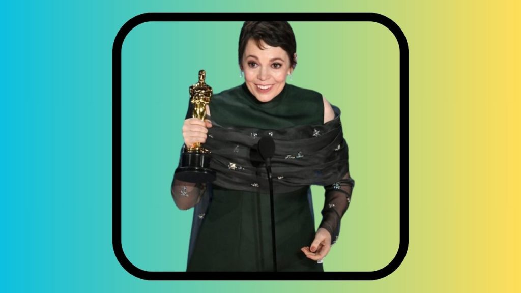 A cropping of a screenshot from the 2019 Oscars, showing Olivia Colman receiving an award for best actress. She is wearing a dark green dress and black sheer shawl and is holding up the trophy.
