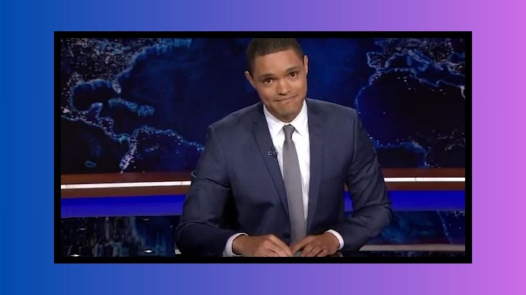 A screenshot from a period of time when Trevor Noah hosted The Daily Show. He is in a crisp dark blue suit.