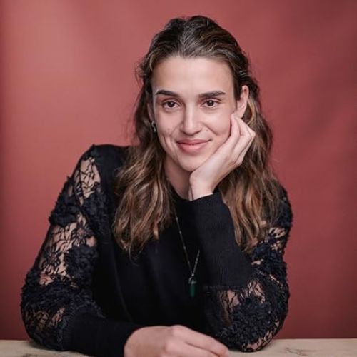 Lydia Goldman, a white woman smiles at the camera while propping her chin and cheek on her hand. She wears a black lace armed shirt before a rust colored background.