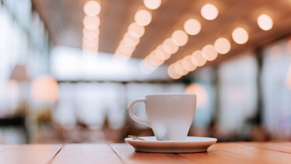 A white coffee mug and saucer sits on a wooden table in a coffee shop.