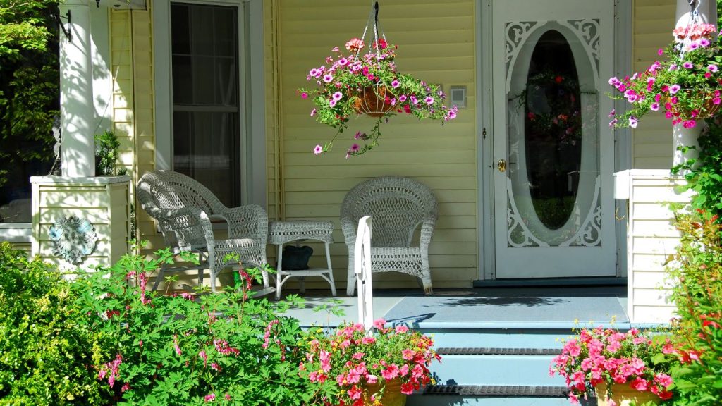 A porch of a yellow house with white wicker chairs and pink flowers and bushes.