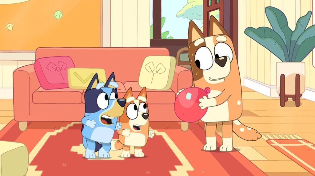 A screenshot from the animated tv show Bluey, featuring an orange adult cartoon dog holding a red balloon in a living room while two child dogs - one blue, one orange - look on with excitement.