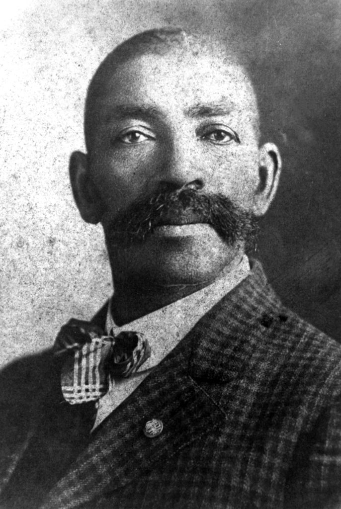A black and white photograph of Black lawman Bass Reeves. He wears a checkered suit and bowtie.