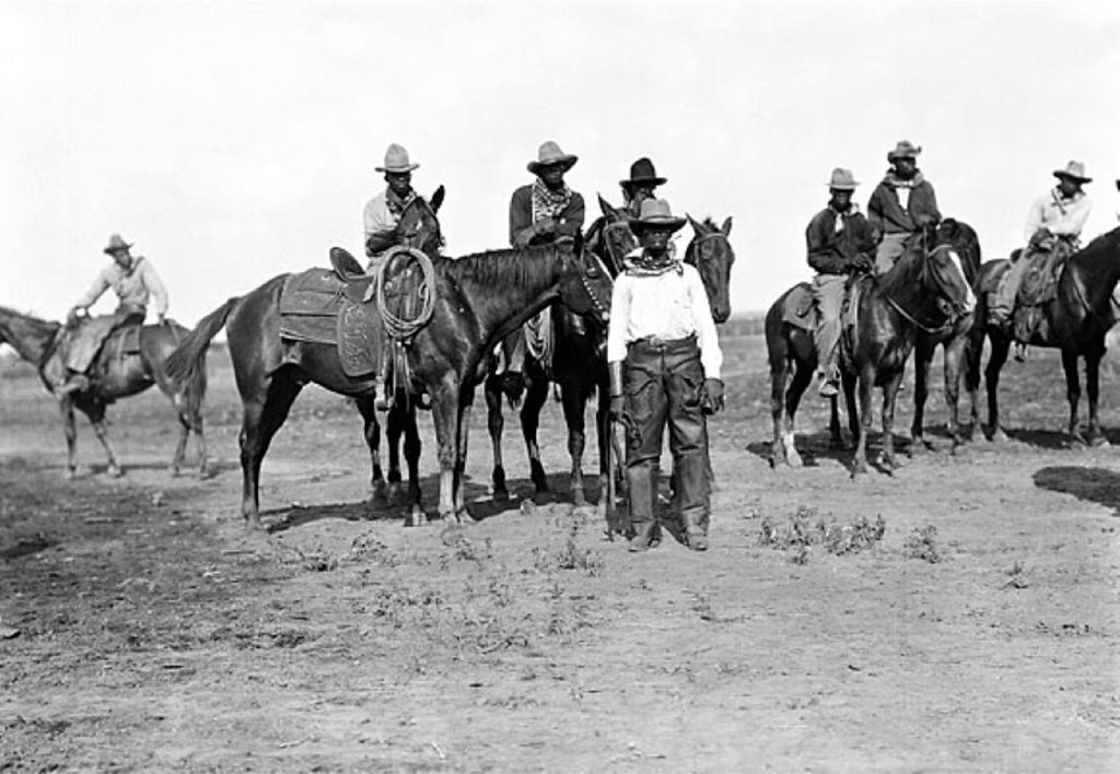A black and white photograph of eight Black cowboys in a barren field. Most are mounted on horses, and one stands in front of the crowd.