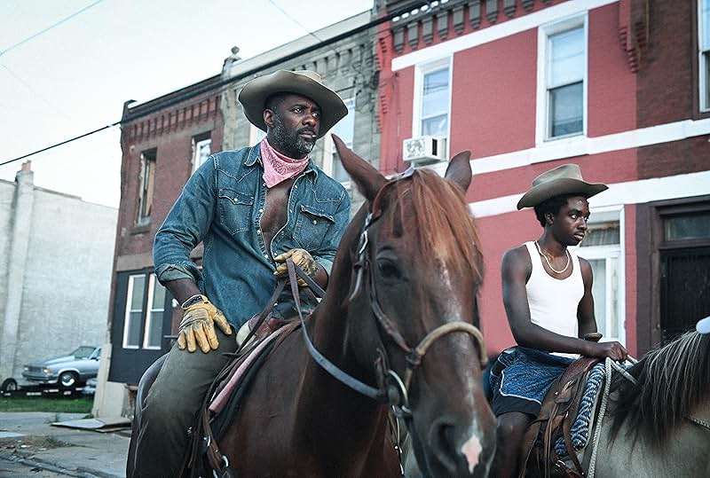 A still from Concrete Cowboy. A Black man and a Black teenage boy ride horses down a city street. The man wears a tan cowboy hat, a denim shirt, a pink bandana, and yellow gloves. The teenager wears a tan cowboy hat and a white tanktop.