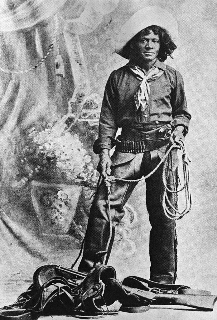A black and white photograph of Black cowboy Nat Love. Love wears a traditional Western cowboy outfit and holds a lasso in his hand. At his feet are a saddle and rifle. Behind him is a drape with painted flower vases on it.