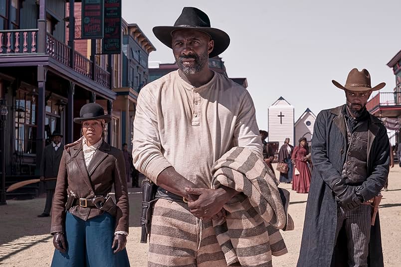 A still from The Harder They Fall. Two Black men and one Black woman stand in the street within a Western town. the woman on the left wears a black bowler hat, a brown jacket, and blue bottoms. The man in the middle wears a black cowboy hat, a white shirt, and carries a prison uniform in his arms. The man on the right wears a brown cowboy hat and a black coat.