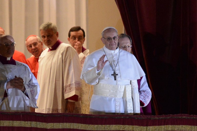 Pope Francis stands on the balcony of St. Peter's Basilica, waving his right hand with a smile. Accompanying him are various officials of the Catholic Church.